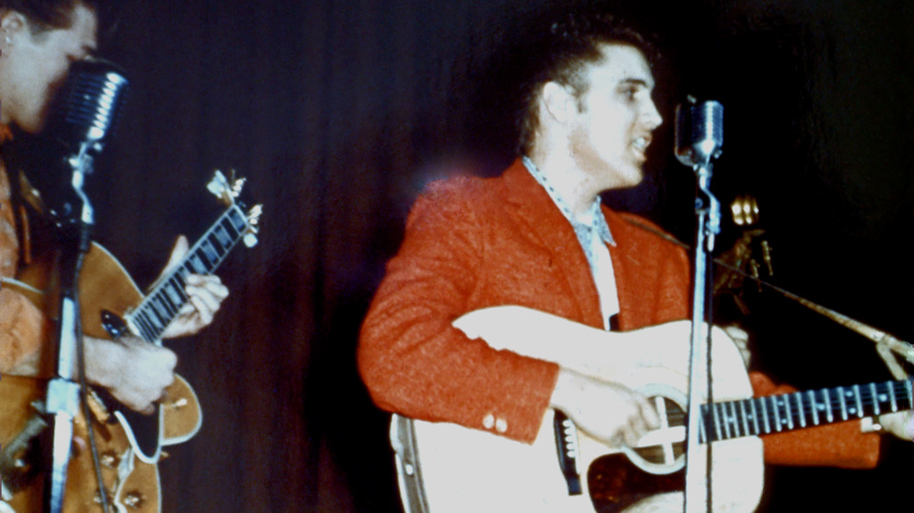 Elvis in a red blazer playing guitar and singing into a mic stand at a high school performance