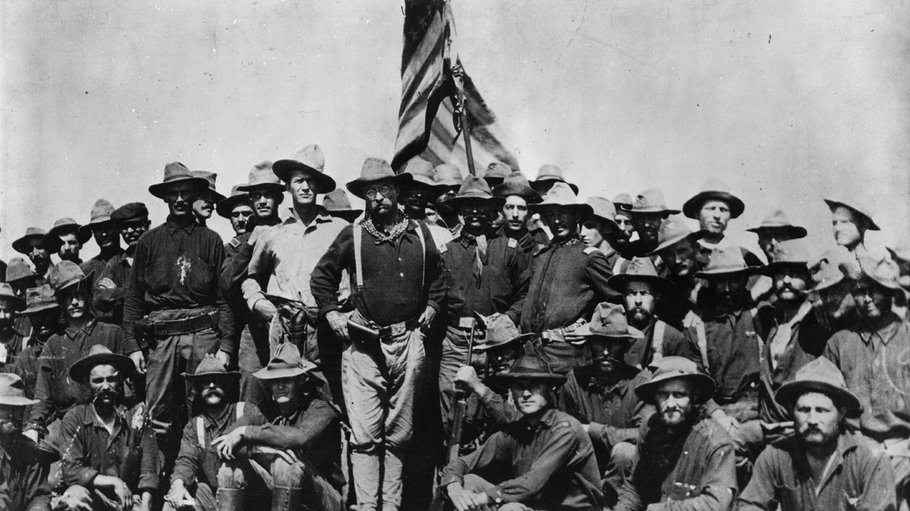 Teddy Roosevelt poses with his Rough Riders in front of flag