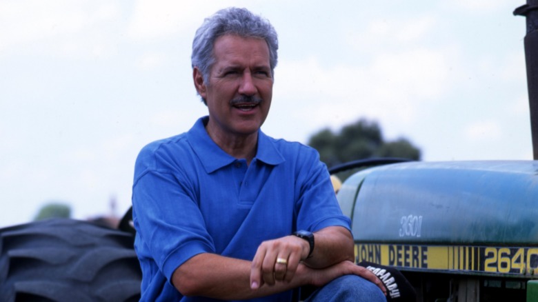 Younger Alex Trebek kneeling on a John Deere tractor