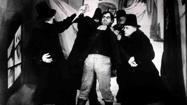 A scene showing men in black with one holding a stake