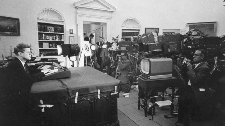 John F. Kennedy behind desk with press watching